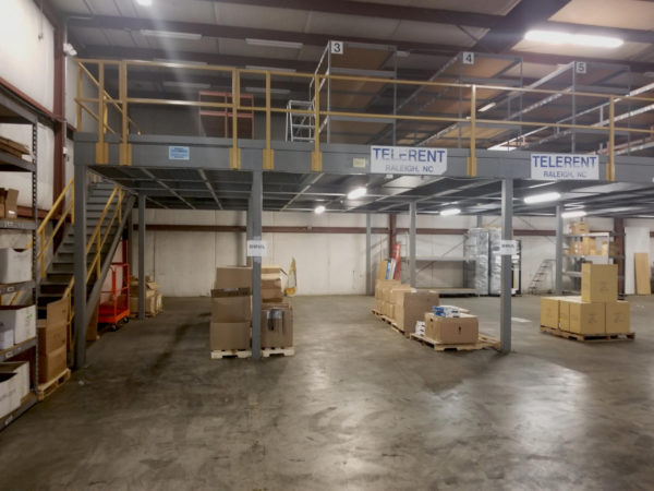 A mezzanine with yellow guard rails set up on gray metal posts, and rivet shelving on the mezzanine.