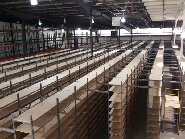 Rivet shelving being set up to form mezzanines, with pallet rack in the background made with particle board shelving, and a gray finish.